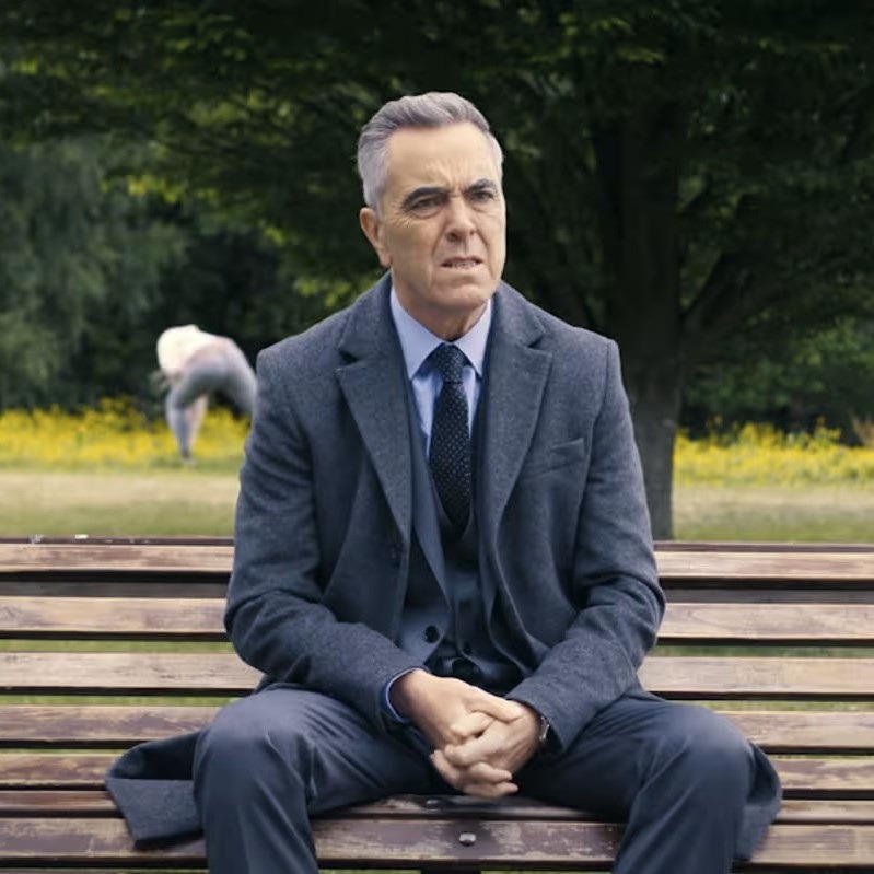 James Nesbitt sits on a wooden bench, wearing a suit, with a park background and blurred figures.