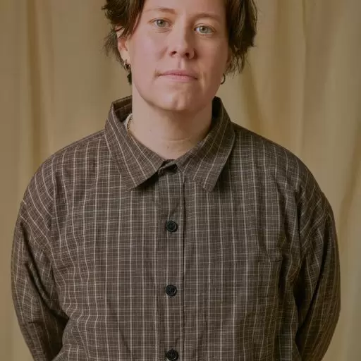 Chloe Petts stands in front of a beige backdrop, wearing a brown checked shirt and looking directly at the camera.