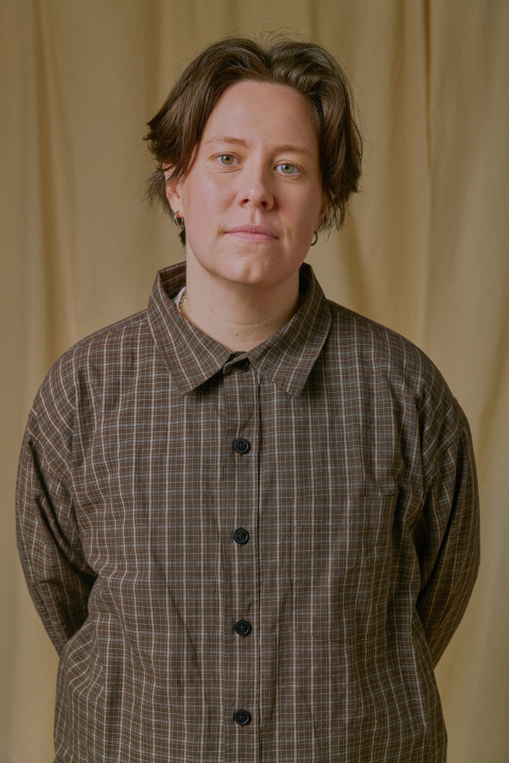 Chloe Petts stands in front of a beige backdrop, wearing a brown checked shirt and looking directly at the camera.
