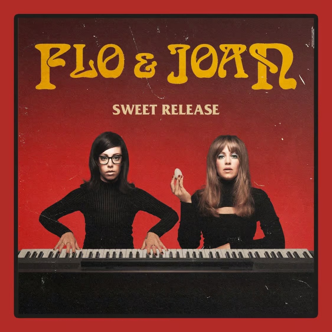 Flo and Joan sit at a keyboard, wearing black outfits, against a red background with yellow text.