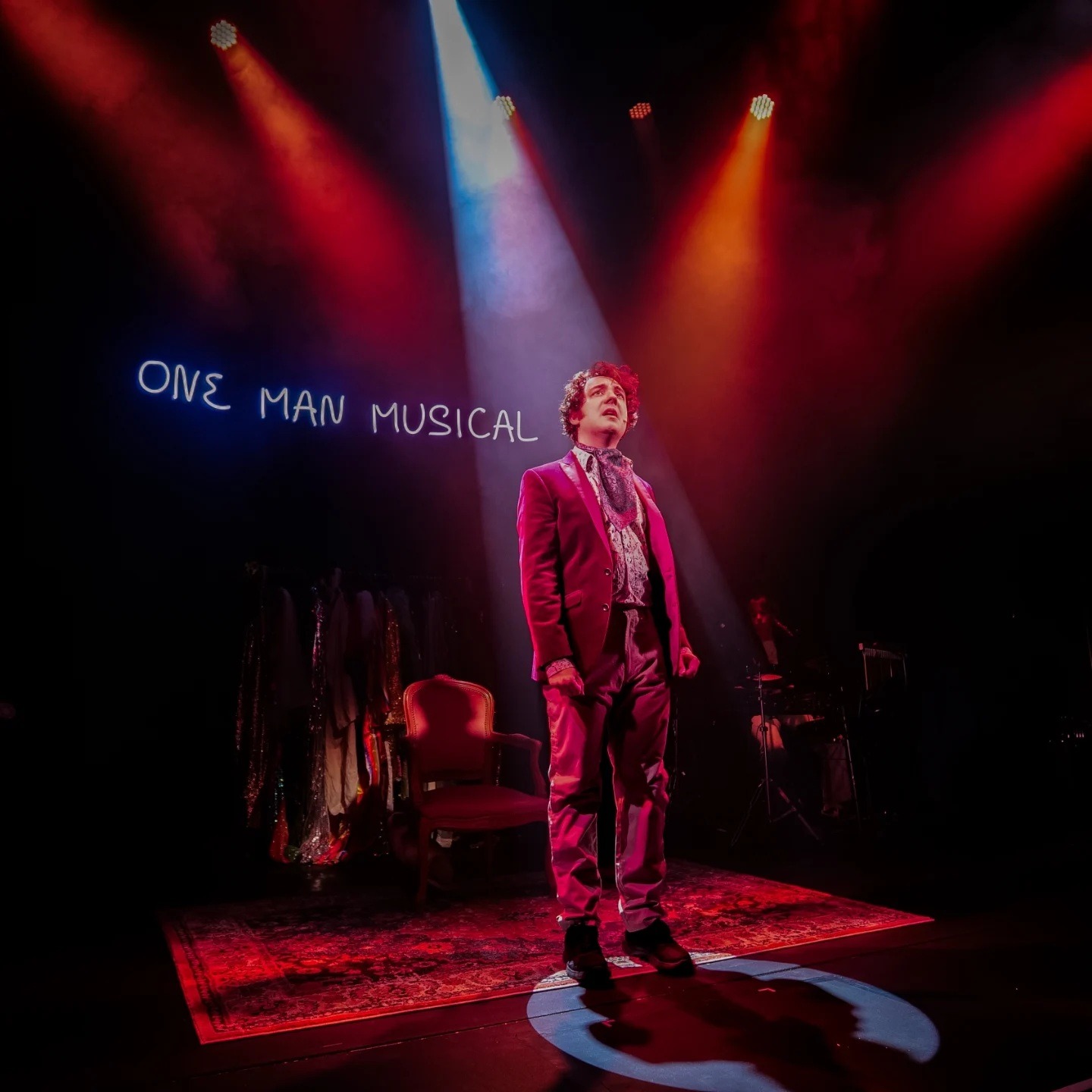 A performer stands centre stage under dramatic lighting, with "One Man Musical" displayed in the background.