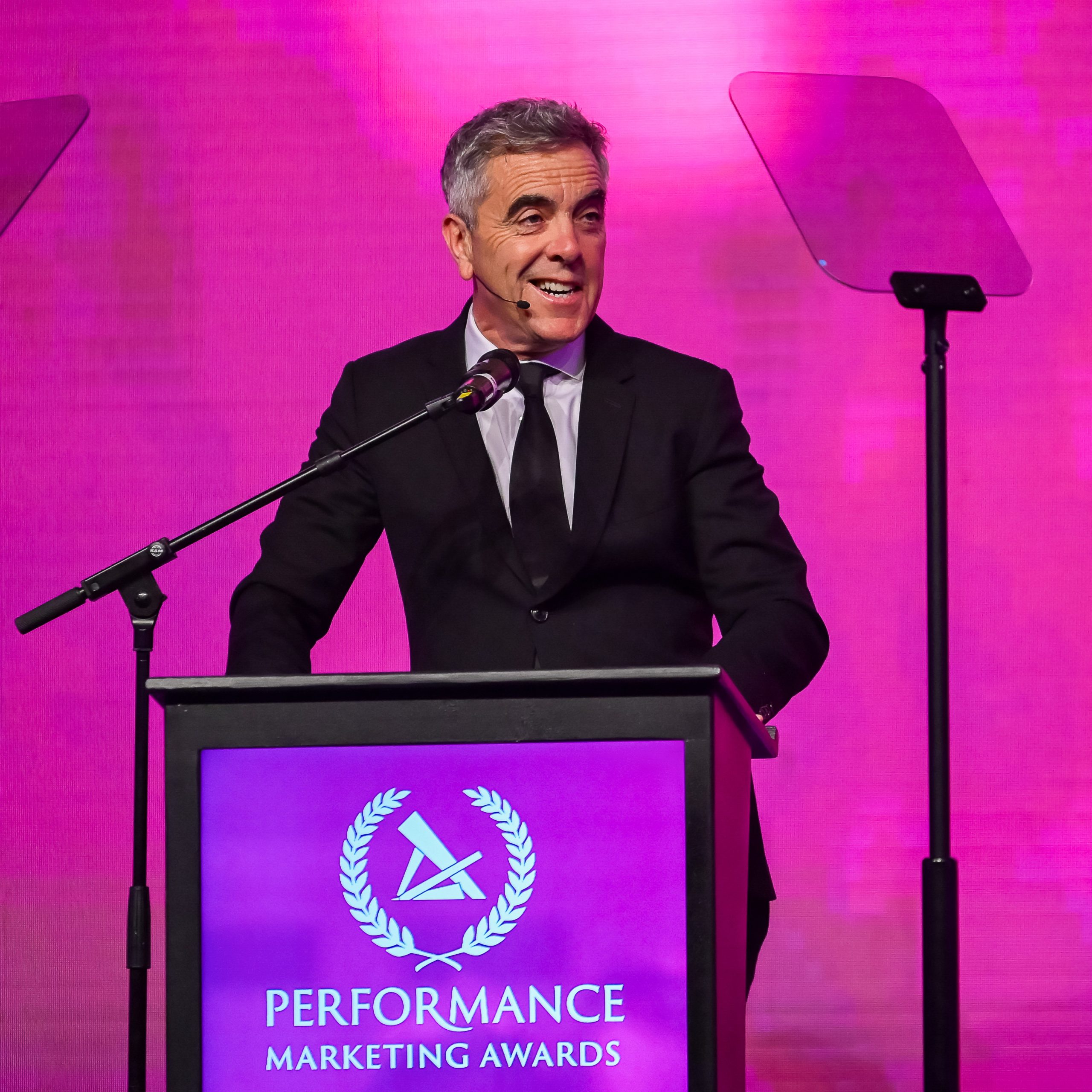 James Nesbitt stands at a podium, smiling, with a pink backdrop and "Performance Marketing Awards" displayed.