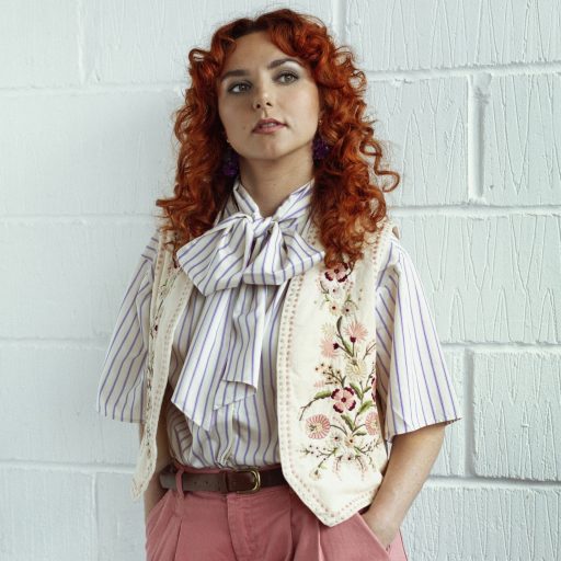 Ania Magliano poses against a white wall, wearing a striped shirt, embroidered vest, and pink trousers.