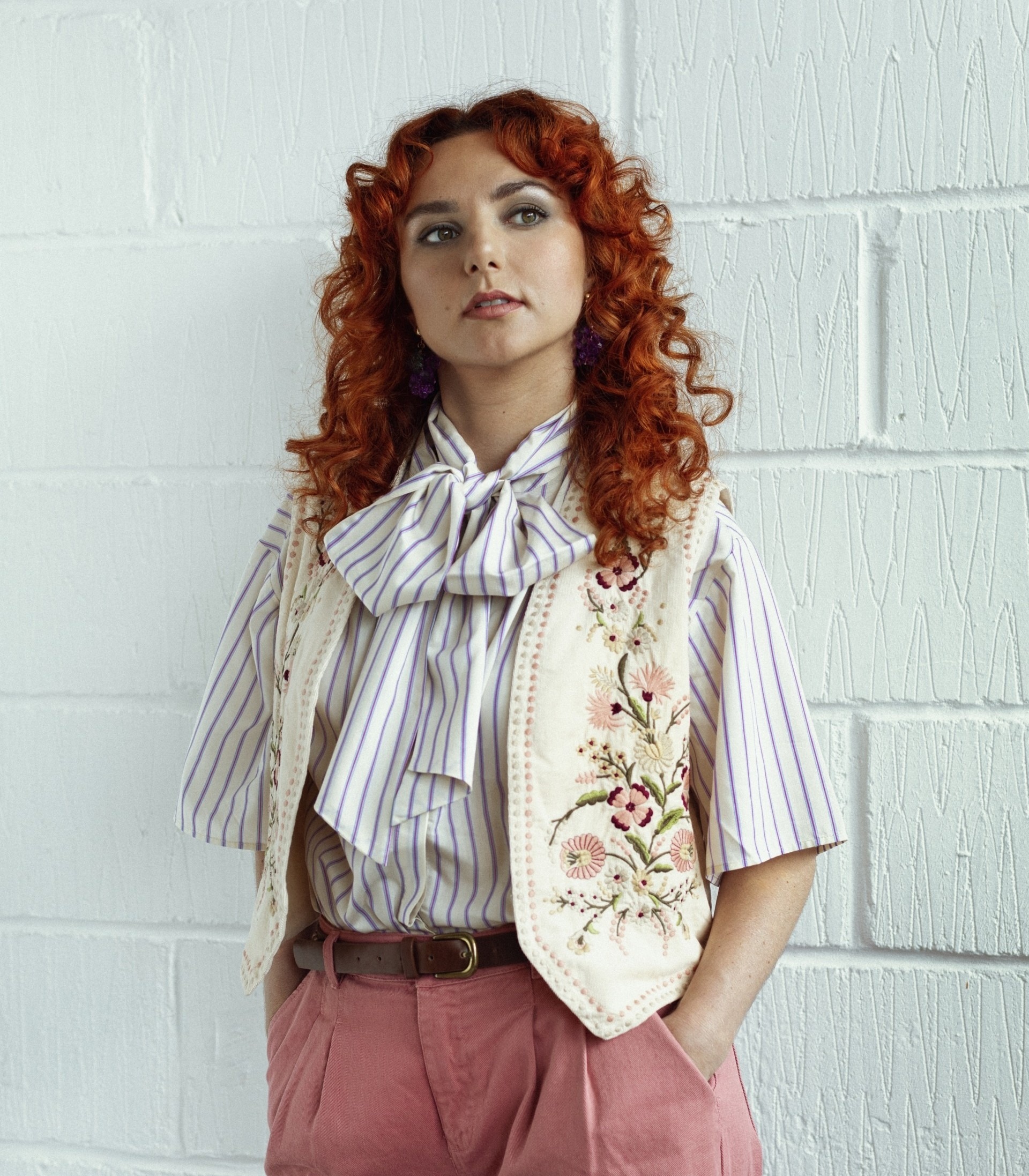 Ania Magliano poses against a white wall, wearing a striped shirt, embroidered vest, and pink trousers.