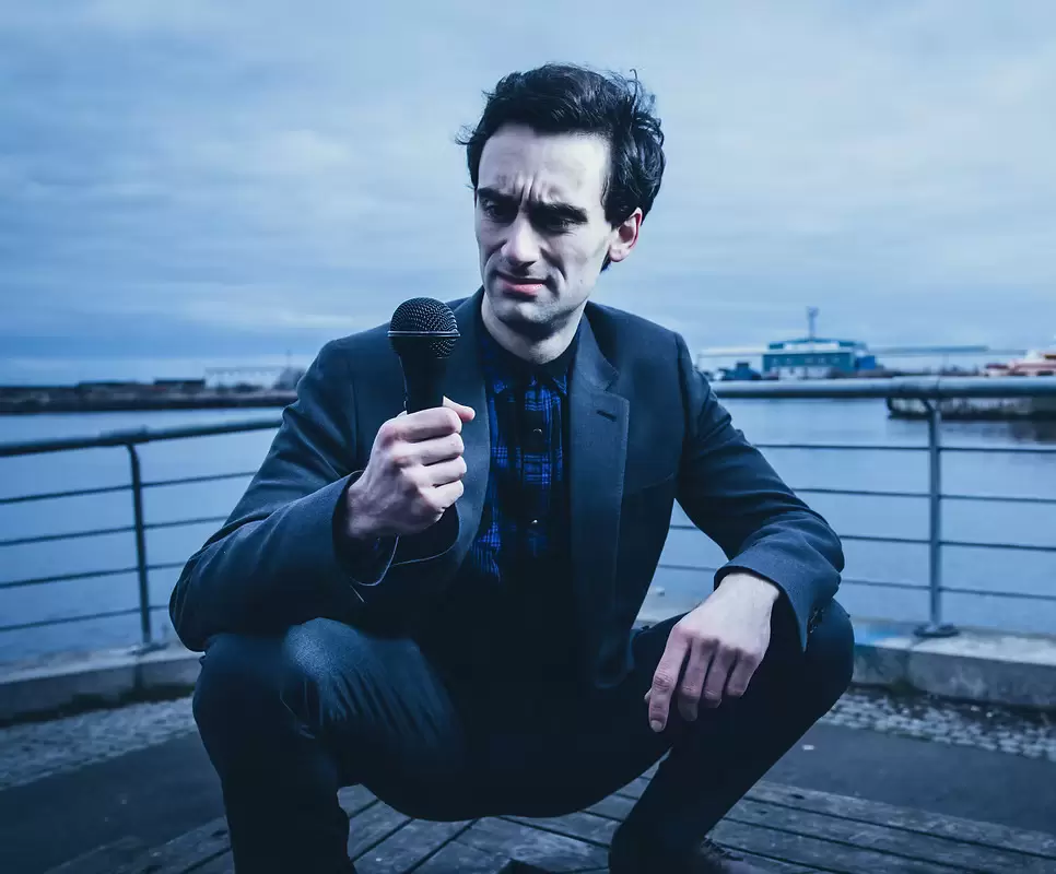 MC Hammersmith squats by a waterfront, holding a microphone, wearing a dark suit and blue shirt.