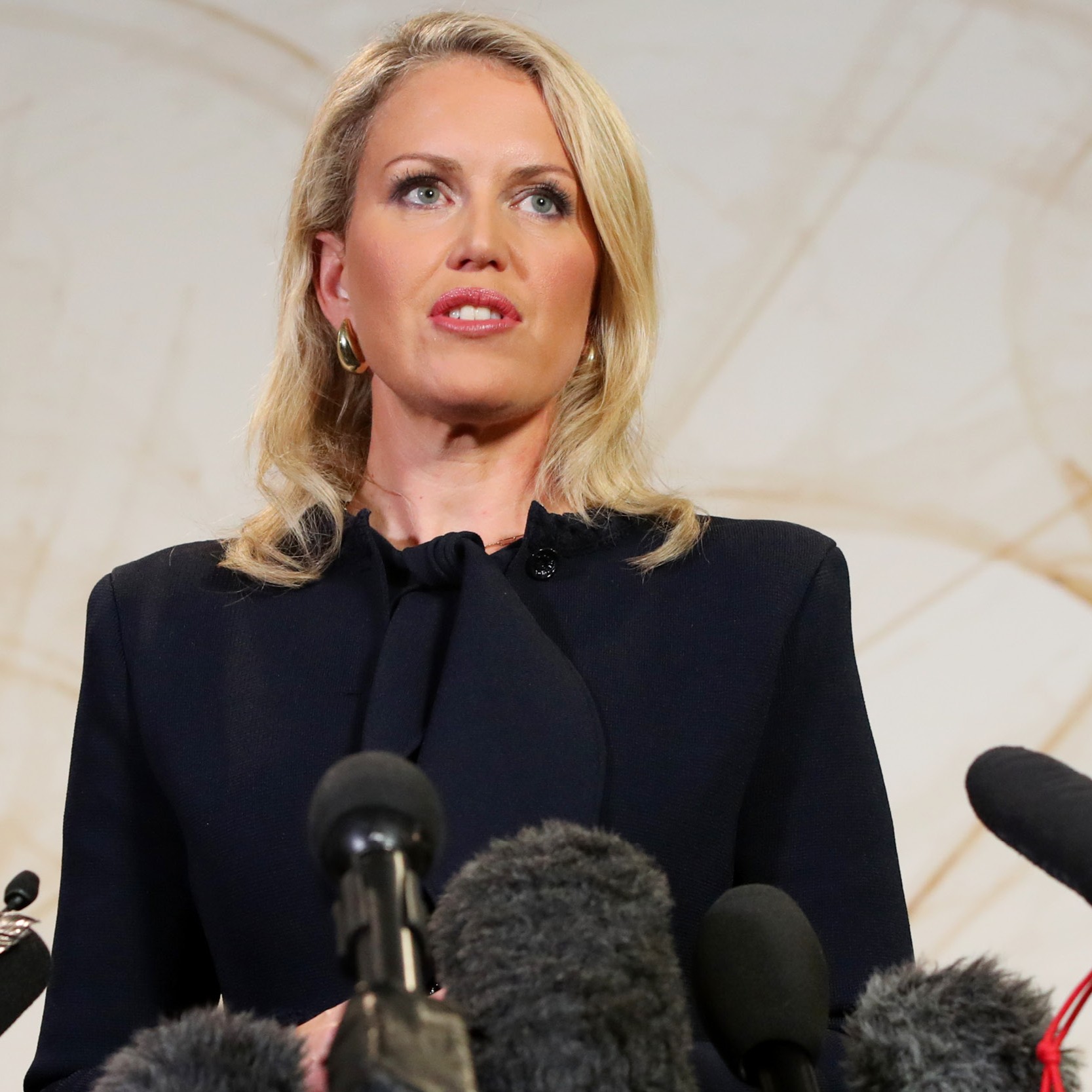 Jennifer Robinson stands at a podium surrounded by microphones, wearing a dark blazer and looking serious.