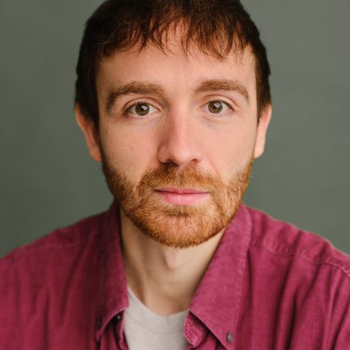 Ian Smith, with reddish-brown hair and beard, poses in a pink shirt against a grey background.